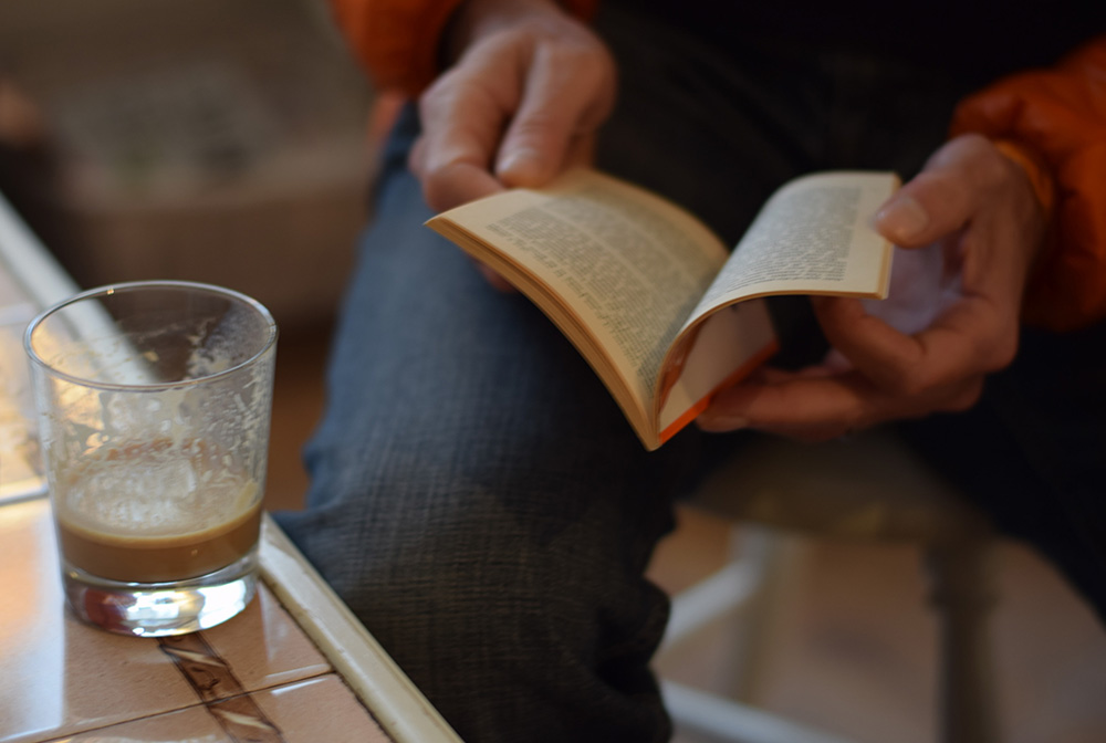 A close up photo of a glass of coffee and a man reading a small book in the Krog of Krinkel Bokcafe in Bergen, Norway