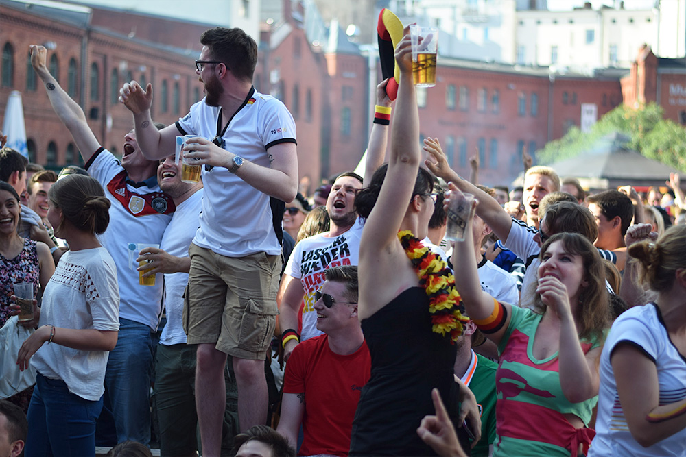 Group of soccer fans celebrating a goal. They are watching a game on a screen in a public square in Berlin, Germany.