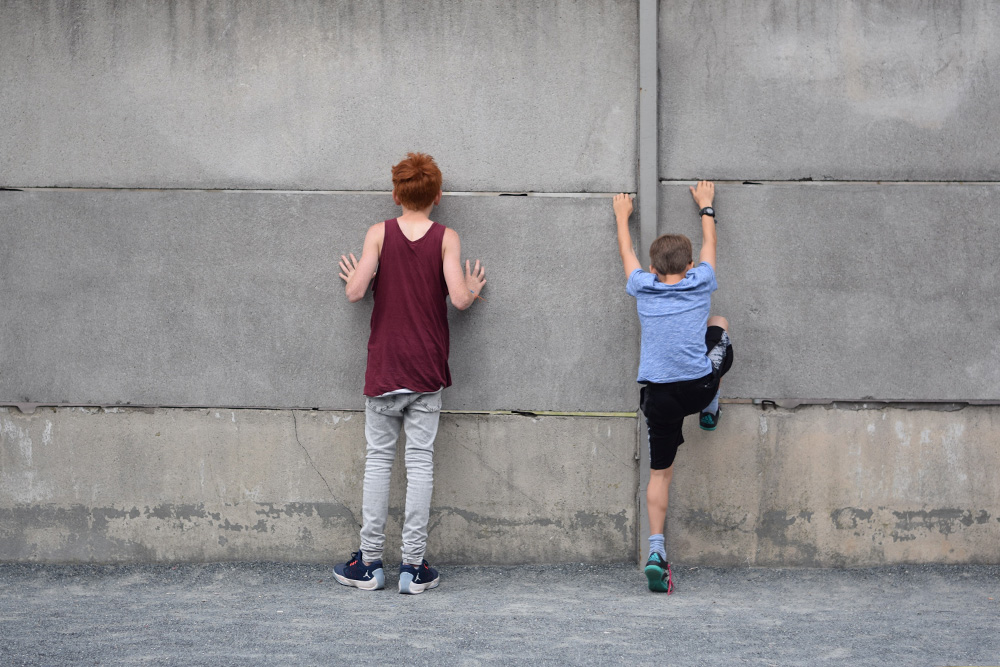 Photo of brothers (tourists) standing at the old Berlin Wall, touching the wall, trying to peer through, one of the boys is trying to climb the wall.