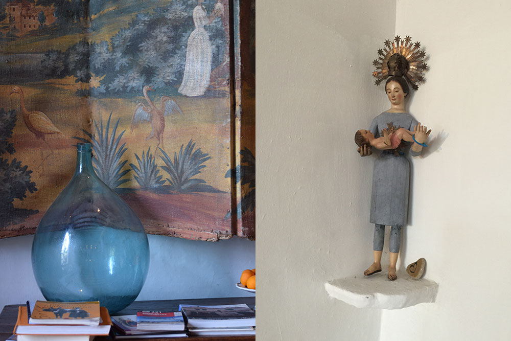 Interior detail of a blue glass bottle on a table with guidebooks, a traditional painting and a religious statue of a women holding a baby.