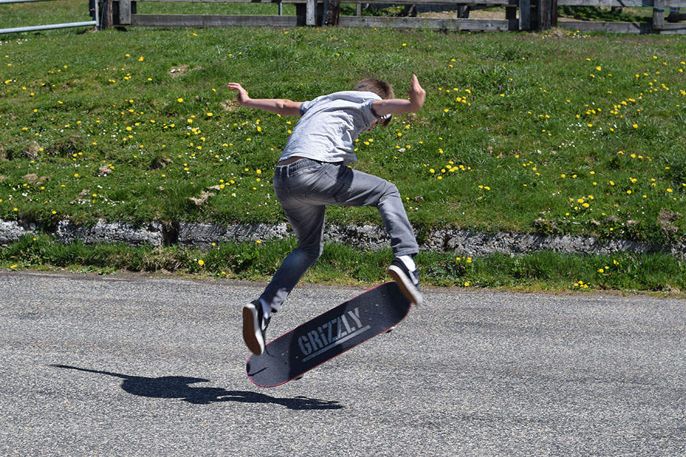 Photo of a boy doing a jumping trick a skateboard with green grass and yellow flowers in the background.