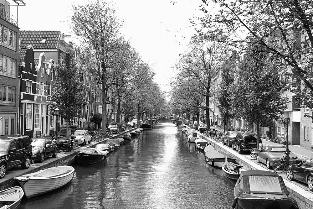 A black and white photo of a canal in Amsterdam lined with boats and cars.